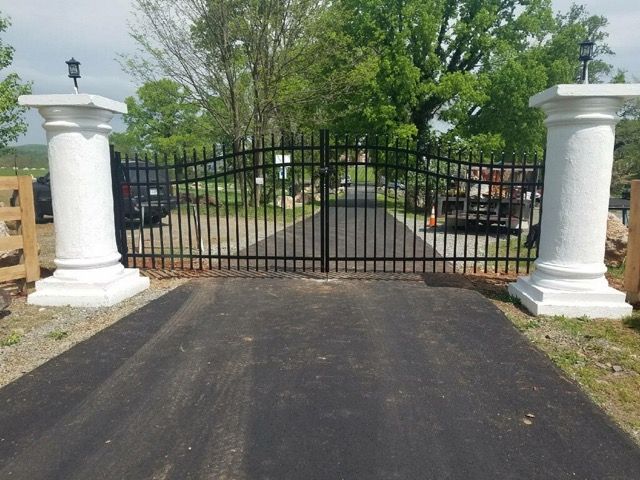 estate-gate-2 A cemetery with a tall fence, gate, and gravestones surrounded by a building, trees, plants, and a bright blue sky outdoors.